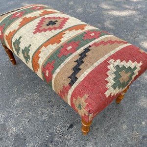 May include: A rectangular ottoman with a woven fabric top featuring a colourful geometric pattern in shades of red, orange, green, brown, and black. The ottoman has wooden legs.