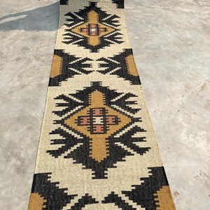 Puede incluir: Una alfombra larga y rectangular con un patr&oacute;n geom&eacute;trico en negro, blanco y marr&oacute;n. La alfombra tiene flecos en los extremos.