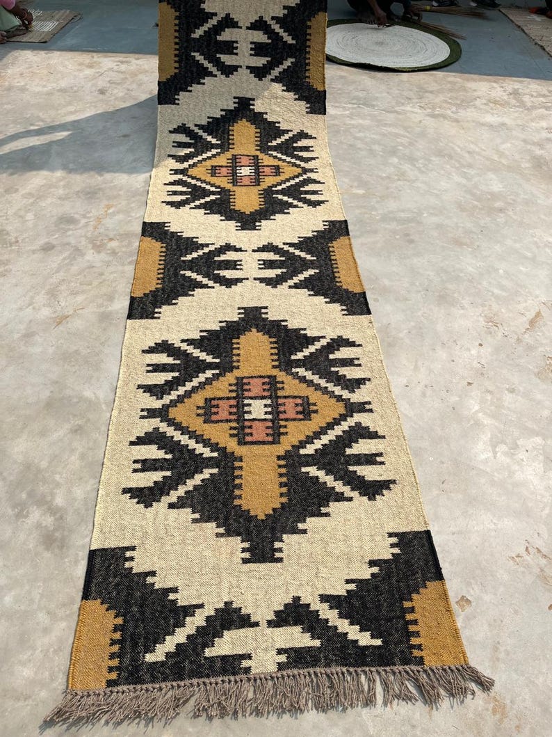 Puede incluir: Una alfombra larga y rectangular con un patr&oacute;n geom&eacute;trico en negro, blanco y marr&oacute;n. La alfombra tiene flecos en los extremos.