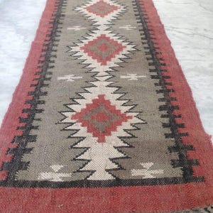 May include: A rectangular rug with a geometric pattern. The rug features a red border and fringe, with a central design in shades of gray, white, and red. The pattern includes diamond and zigzag shapes.