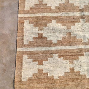 May include: A brown and white woven rug with a geometric pattern. The rug has a natural, rustic look and is made of jute.