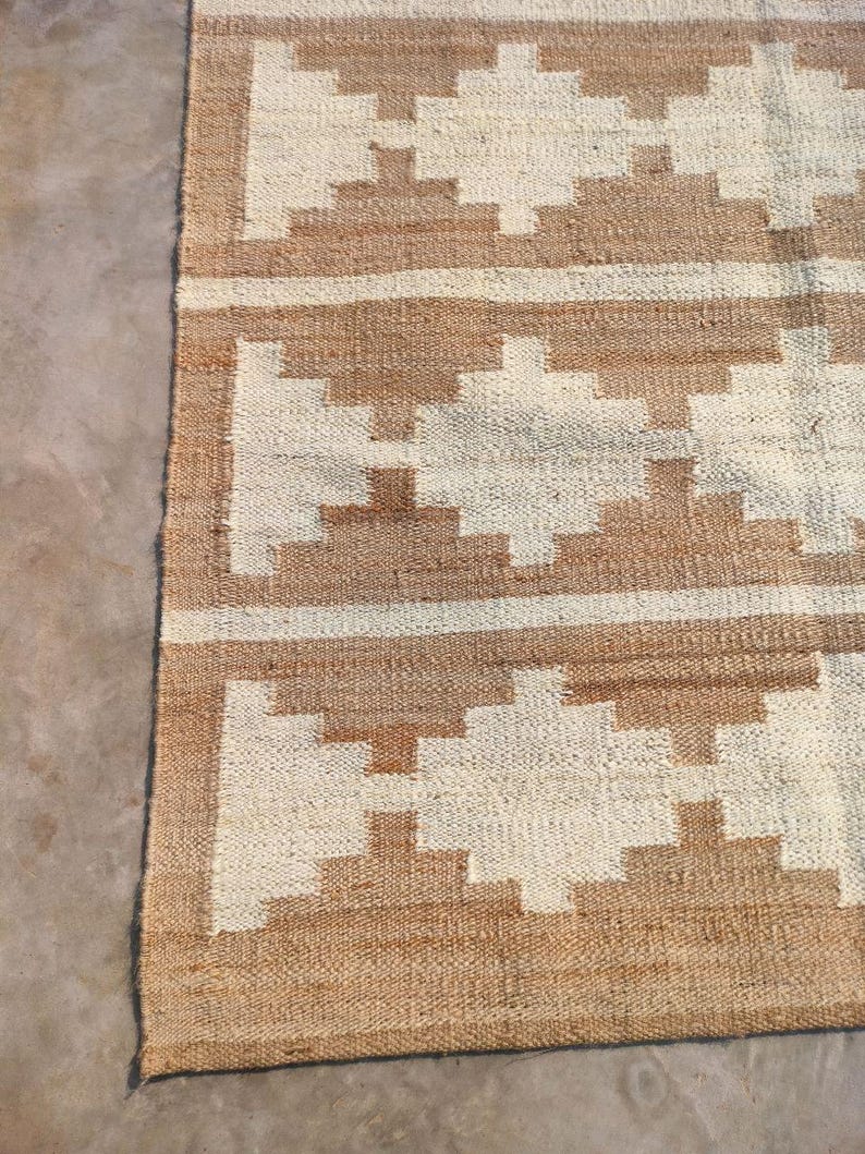 May include: A brown and white woven rug with a geometric pattern. The rug has a natural, rustic look and is made of jute.