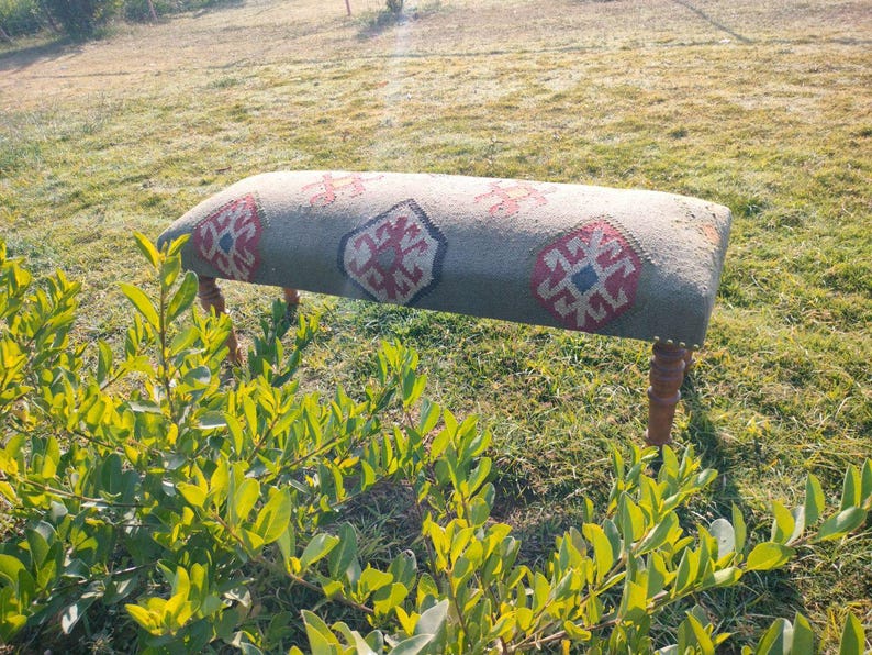 Op de afbeelding: Een houten bank met een kleurrijk, gepatroneerd kussen. Het kussen heeft een geometrisch ontwerp met rode, blauwe en witte accenten. De bank staat op een grasveld.