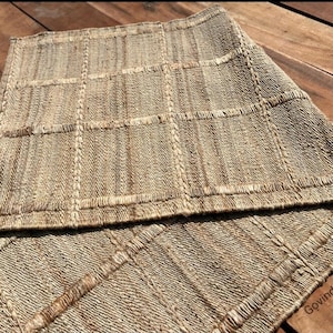 May include: A woven rug made of natural fibers with a checkered pattern. The rug is light brown and beige in color. The rug is folded in half and the brand name "Govind Art Rugs" is visible in the bottom right corner.