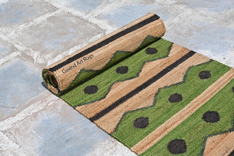 May include: A rolled-up jute rug with a green and tan geometric pattern. The rug features black accents and circular designs. The text "Govind Art Rugs" is visible on the rolled portion of the rug.