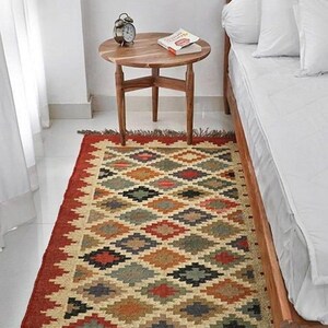 May include: A rectangular area rug with a geometric diamond pattern in red, beige, green, and brown. The rug has a red border and fringe. A wooden side table with a book and clock sits nearby.
