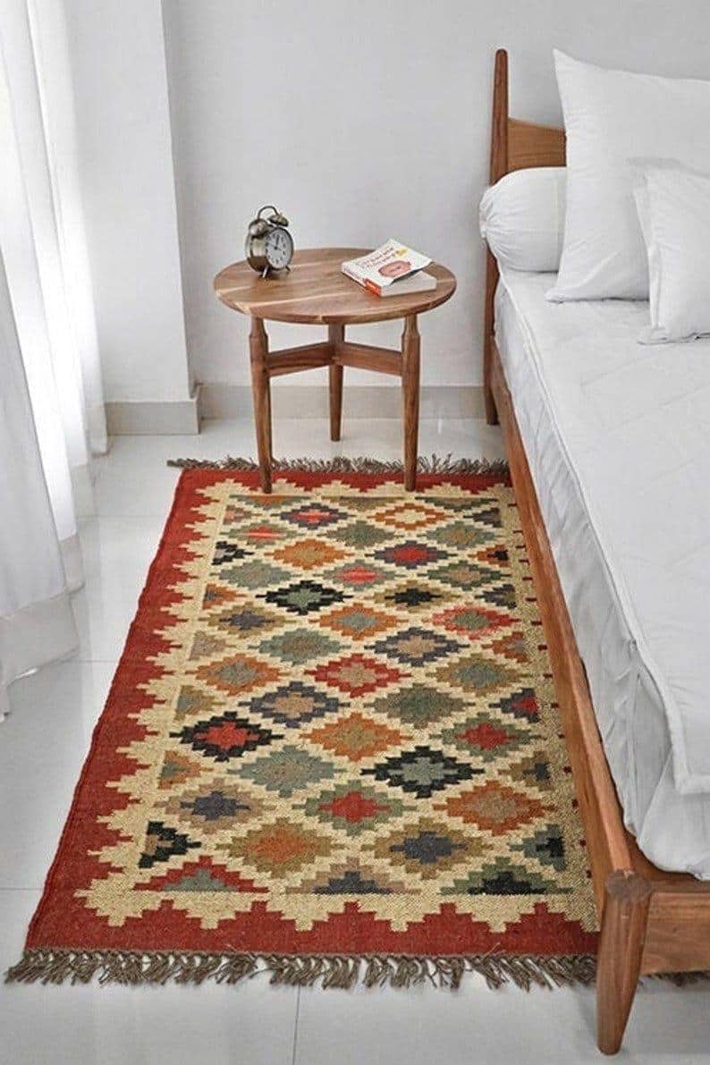 May include: A rectangular area rug with a geometric diamond pattern in red, beige, green, and brown. The rug has a red border and fringe. A wooden side table with a book and clock sits nearby.