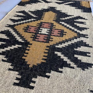 Puede incluir: Una alfombra de color beige, negro y marr&oacute;n con un dise&ntilde;o geom&eacute;trico. La alfombra tiene una textura tejida y est&aacute; hecha de fibras naturales.