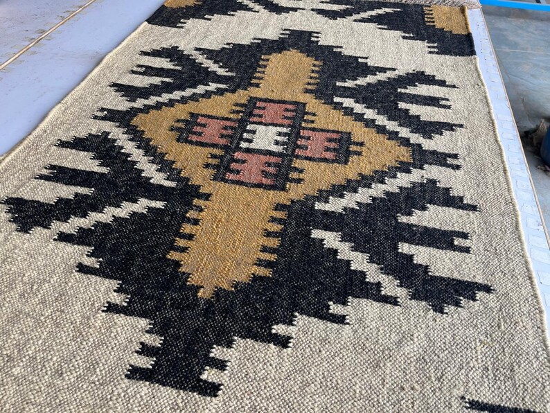 Puede incluir: Una alfombra de color beige, negro y marr&oacute;n con un dise&ntilde;o geom&eacute;trico. La alfombra tiene una textura tejida y est&aacute; hecha de fibras naturales.