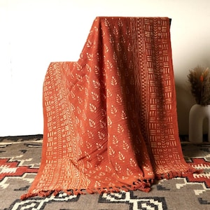 May include: A rust-colored throw blanket with a white leaf pattern and geometric design. The blanket has fringe on the bottom edge.