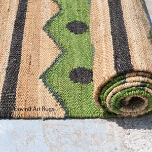 May include: A rolled-up jute rug with a geometric design. The rug features a natural tan background with green and black accents. The design includes zigzags, stripes, and circular shapes. The text "Govind Art Rugs" is visible.