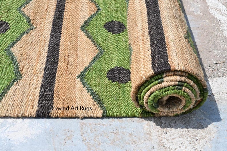 May include: A rolled-up jute rug with a geometric design. The rug features a natural tan background with green and black accents. The design includes zigzags, stripes, and circular shapes. The text "Govind Art Rugs" is visible.