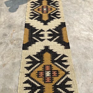 Puede incluir: Una alfombra rectangular larga con un patr&oacute;n geom&eacute;trico en tonos beige, marr&oacute;n y negro. La alfombra tiene un borde con flecos.