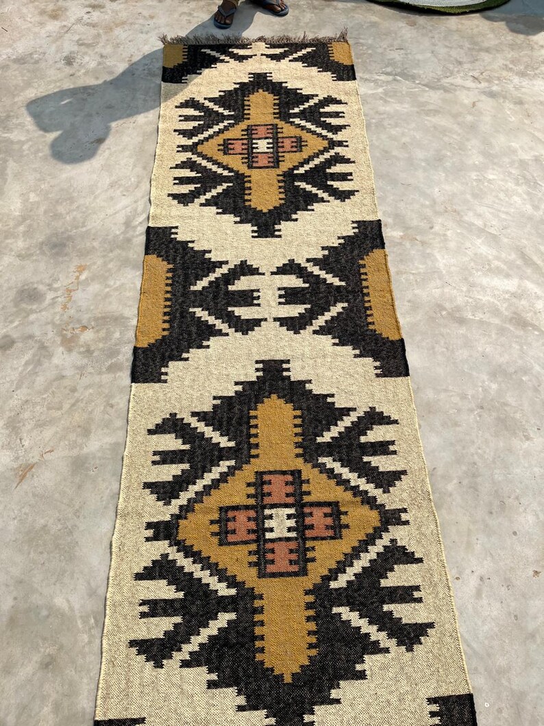 Puede incluir: Una alfombra rectangular larga con un patr&oacute;n geom&eacute;trico en tonos beige, marr&oacute;n y negro. La alfombra tiene un borde con flecos.