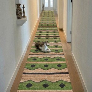 May include: A long, green and beige runner rug with a geometric pattern, laid out in a hallway. The rug features a repeating design of green zigzags and black dots. A cat is lying on the rug. The text "Govind Art Rugs" is at the bottom.