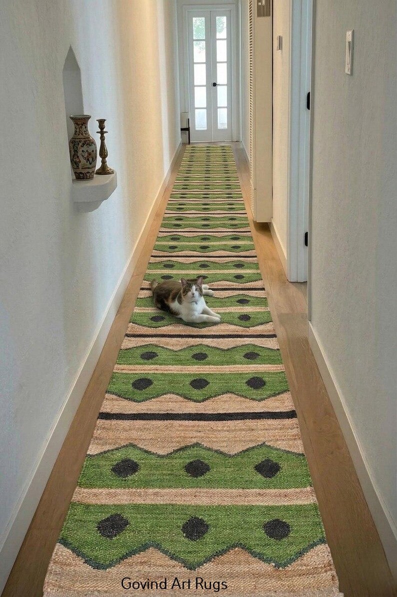 May include: A long, green and beige runner rug with a geometric pattern, laid out in a hallway. The rug features a repeating design of green zigzags and black dots. A cat is lying on the rug. The text "Govind Art Rugs" is at the bottom.