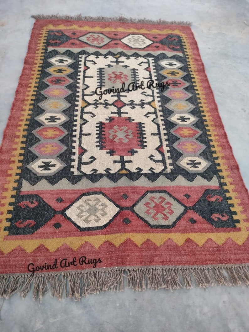 May include: A hand-woven rug with a geometric pattern in shades of red, black, white, and beige. The rug has a fringe border and the text "Govind Art Rugs" is visible in the bottom right corner.