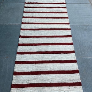 Op de afbeelding: Een wit en bordeaux gestreept tapijt met de tekst "Govind Art Rugs" onderaan.
