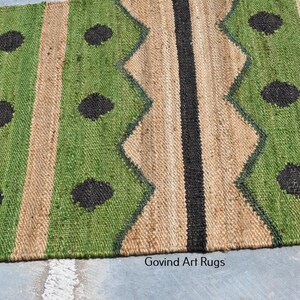 May include: A jute rug with a geometric pattern. The rug features alternating vertical stripes of green, tan, and black. The green stripes have a zig-zag design and black circular accents. The rug is made of natural fibers.
