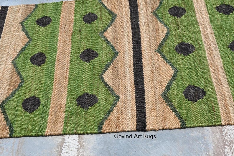 May include: A jute rug with a geometric pattern. The rug features alternating vertical stripes of green, tan, and black. The green stripes have a zig-zag design and black circular accents. The rug is made of natural fibers.
