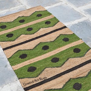 May include: A rectangular jute rug with a repeating pattern of green and black shapes on a tan background. The rug has a geometric design with wavy green stripes and black circles. The words "Govind Art Rugs" are visible.