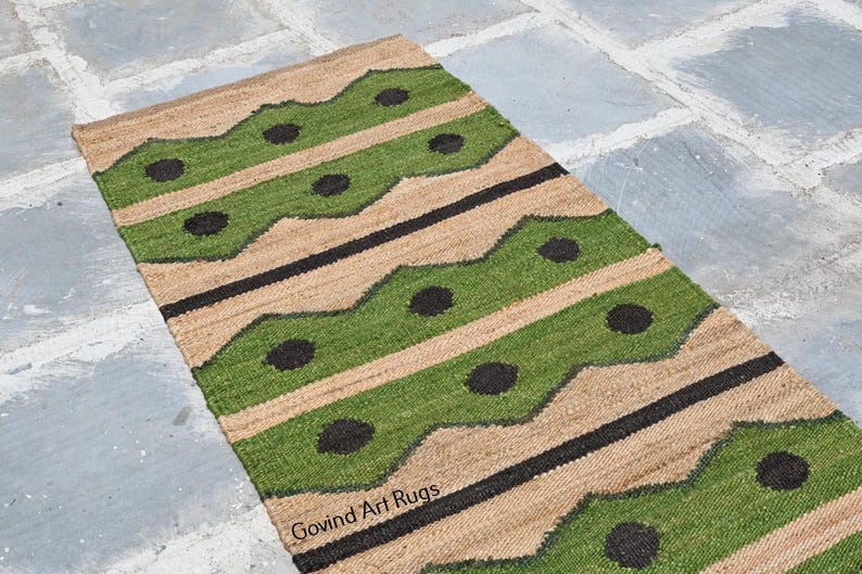 May include: A rectangular jute rug with a repeating pattern of green and black shapes on a tan background. The rug has a geometric design with wavy green stripes and black circles. The words "Govind Art Rugs" are visible.