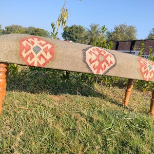 Op de afbeelding: Een houten bank met een geweven tapijt bovenop met een geometrisch patroon in tinten rood, blauw en wit. De bank heeft vier houten poten.