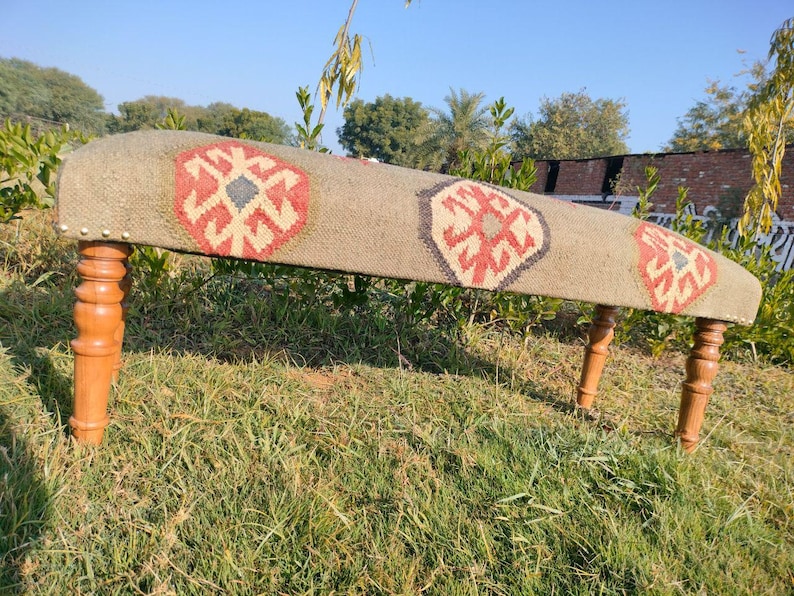 Op de afbeelding: Een houten bank met een geweven tapijt bovenop met een geometrisch patroon in tinten rood, blauw en wit. De bank heeft vier houten poten.