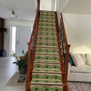 May include: A staircase with a green and tan patterned runner rug. The rug features a repeating geometric design with black accents. The wooden stairs have a dark finish, and the walls are painted white. The text "Govind Art Rugs" is visible.