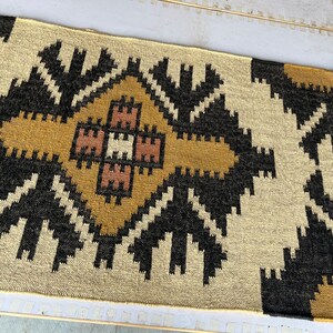 Puede incluir: Una alfombra tejida beige y marr&oacute;n con un patr&oacute;n geom&eacute;trico. La alfombra tiene una forma de diamante central con un diamante m&aacute;s peque&ntilde;o en su interior. La alfombra tiene un borde de flecos.
