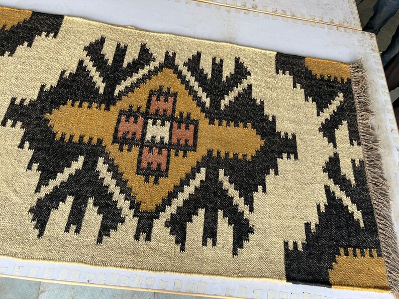 Puede incluir: Una alfombra tejida beige y marr&oacute;n con un patr&oacute;n geom&eacute;trico. La alfombra tiene una forma de diamante central con un diamante m&aacute;s peque&ntilde;o en su interior. La alfombra tiene un borde de flecos.