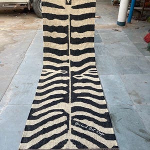 Puede incluir: Una alfombra larga y rectangular con un estampado de rayas de cebra en negro y beige. La alfombra tiene un borde con flecos y el texto "Govind Art Rugs" en la esquina inferior derecha. La alfombra está hecha de un material tejido.