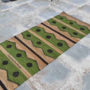 May include: A rectangular runner rug with a geometric pattern. The rug features alternating stripes of green, tan, and black. The green sections have black circular accents. The text "Govind Art Rugs" is visible on the rug.