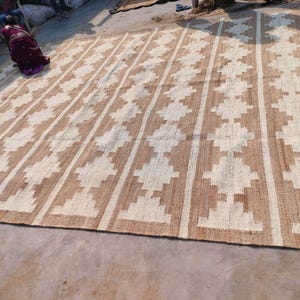 May include: A large, rectangular, woven rug with a geometric pattern. The rug is made of natural fibres and has a light brown and white colour scheme.