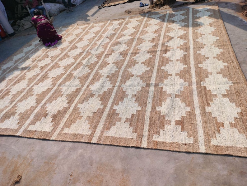 May include: A large, rectangular, woven rug with a geometric pattern. The rug is made of natural fibres and has a light brown and white colour scheme.