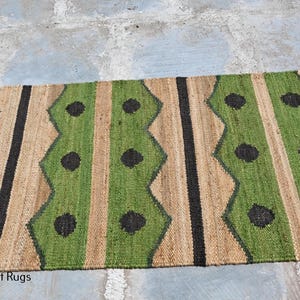 May include: A rectangular jute rug with a repeating pattern of green, tan, and black vertical stripes. The green stripes have a zig-zag design with black circular accents. The rug is suitable for indoor use.