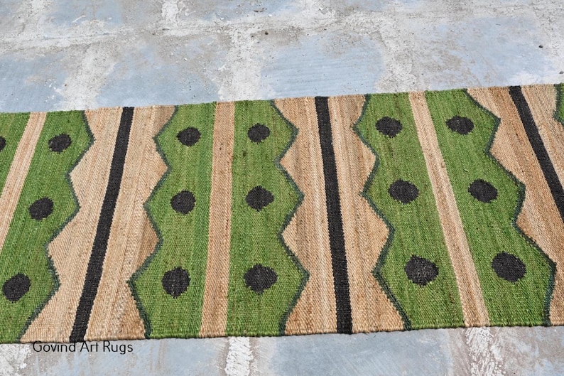 May include: A rectangular jute rug with a repeating pattern of green, tan, and black vertical stripes. The green stripes have a zig-zag design with black circular accents. The rug is suitable for indoor use.