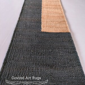 May include: A long, rectangular rug with a dark green and natural brown color scheme. The rug is made of woven fibers and has a textured surface. The text "Govind Art Rugs" is printed in white on the rug.