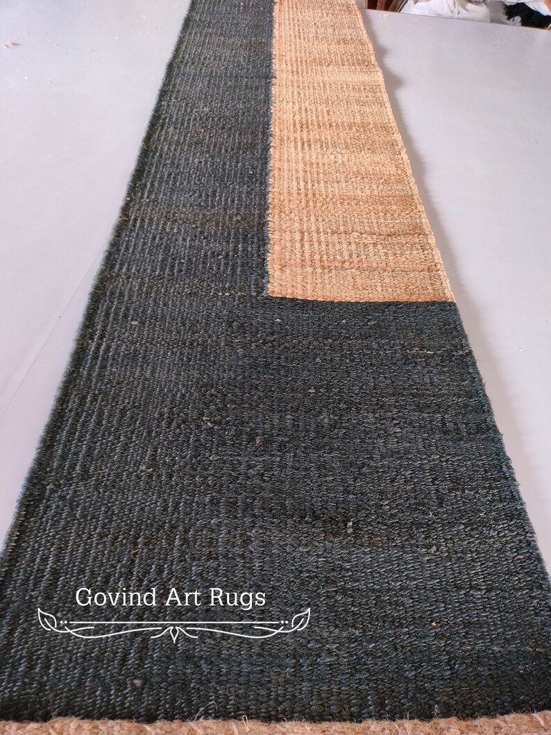 May include: A long, rectangular rug with a dark green and natural brown color scheme. The rug is made of woven fibers and has a textured surface. The text "Govind Art Rugs" is printed in white on the rug.