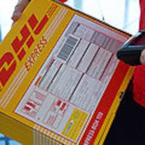 May include: A yellow DHL Express box with red lettering and a barcode is being scanned. The box is held by a person wearing a red jacket. The box is labeled "EXPRESS BOX 120".