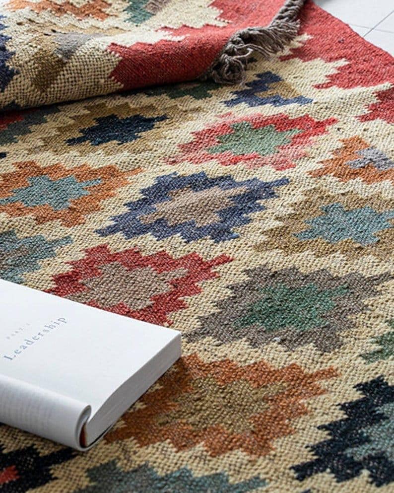 May include: A colorful area rug with a geometric pattern in shades of red, blue, green, and brown. The rug has a textured appearance and a rolled-up section revealing a fringe edge. A book with the word "Leadership" is on the rug.