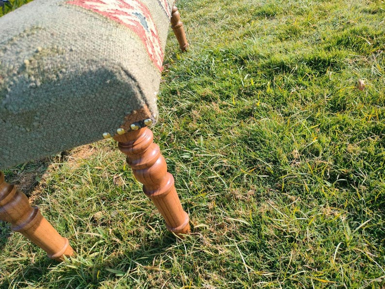 Op de afbeelding: Een houten voetenbankje met een grijze en rode stoffen bovenkant. De voetenbank heeft gedraaide houten poten.