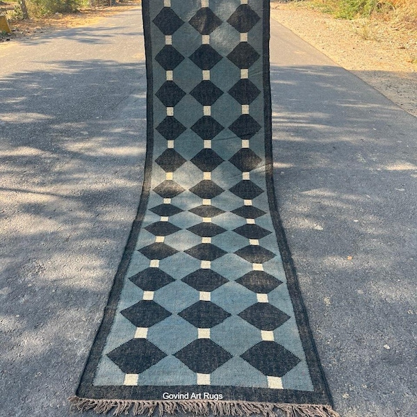 May include: A long, rectangular rug with a geometric diamond pattern in shades of blue and black, accented with white squares. The rug has a fringed edge and is displayed outdoors. The text "Govind Art Rugs" is visible.