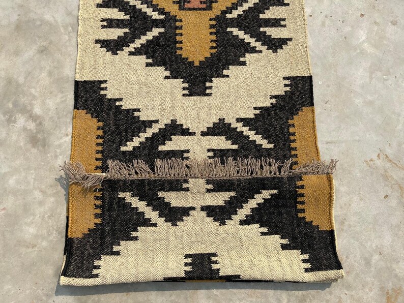 Puede incluir: Una alfombra con estampado geom&eacute;trico en blanco y negro con un borde amarillo mostaza. La alfombra tiene flecos en los bordes.
