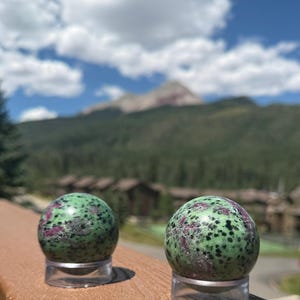 May include: Two polished spheres made of zoisite with ruby inclusions, featuring a mottled green base with dark black speckles and vibrant magenta-red patches. Each stone sphere is displayed resting on a small, clear, circular acrylic stand.