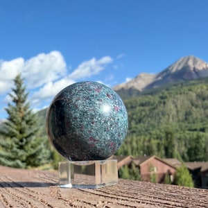 May include: A polished spherical stone featuring a deep teal and dark green base with scattered, vibrant magenta-red inclusions, resting on a clear, square acrylic stand that provides a stable base for the smooth, rounded mineral object.