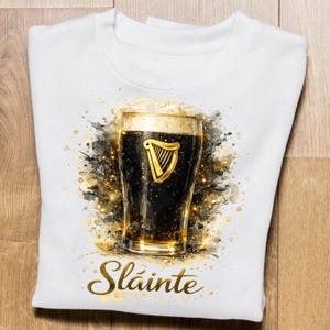 May include: White sweatshirt featuring a watercolor-style image of a pint of dark beer with a golden harp logo. The word "Sl&aacute;inte" is written in gold script below the pint. The design has gold and black accents.