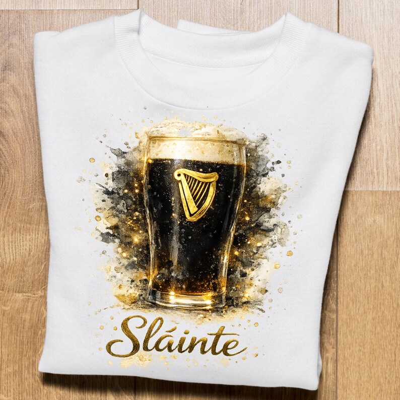 May include: White sweatshirt featuring a watercolor-style image of a pint of dark beer with a golden harp logo. The word "Sl&aacute;inte" is written in gold script below the pint. The design has gold and black accents.