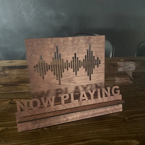 Op de afbeelding: Een houten bord met een geluidgolfontwerp en de woorden "NOW PLAYING" in een uitgesneden lettertype. Het bord is bruin en heeft een plank eronder.