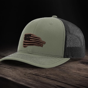 Könnte beinhalten: Eine grüne und schwarze Trucker-Kappe mit einem abgenutzten amerikanischen Flaggen-Patch auf der Vorderseite.